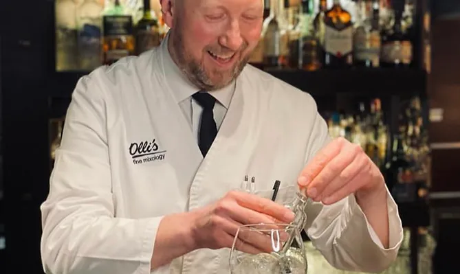 Ein fröhlicher Mann in weißem Kittel mischt Cocktails hinter einer Theke vor einem gut bestückten Rückbuffet.