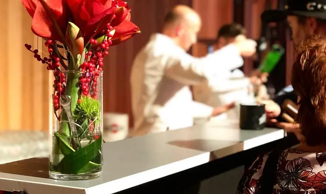 Blick auf Bar, Theke mit Besucherin, die ein geblümtes Kleid und einen Hut trägt, im Hintergrund zwei Mitarbeiter der Bar, vermutlich bei der Zubereitung eines Cocktails. Die Bildmitte ist eine Vase mit roten Lilien.