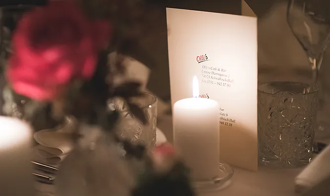 Auf einem Tisch mit einer roten Rose und Kristallgläsern steht hinter einer Kerze von dieser beschienen die Speisekarte vom Olli's